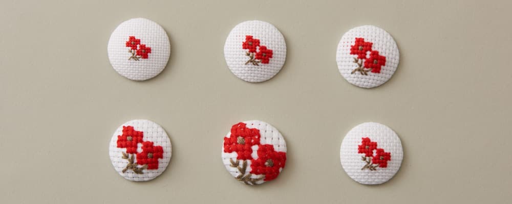 The same floral cross-stitch motif worked on six embroidery cloth counts, shown on covered buttons