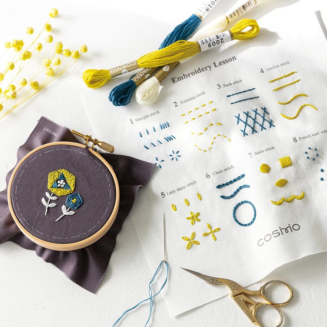 COSMO Embroidery Lesson flat lay with stitch sampler cloth, geometric flower hoop, cream teal and yellow floss, needle, and gold scissors