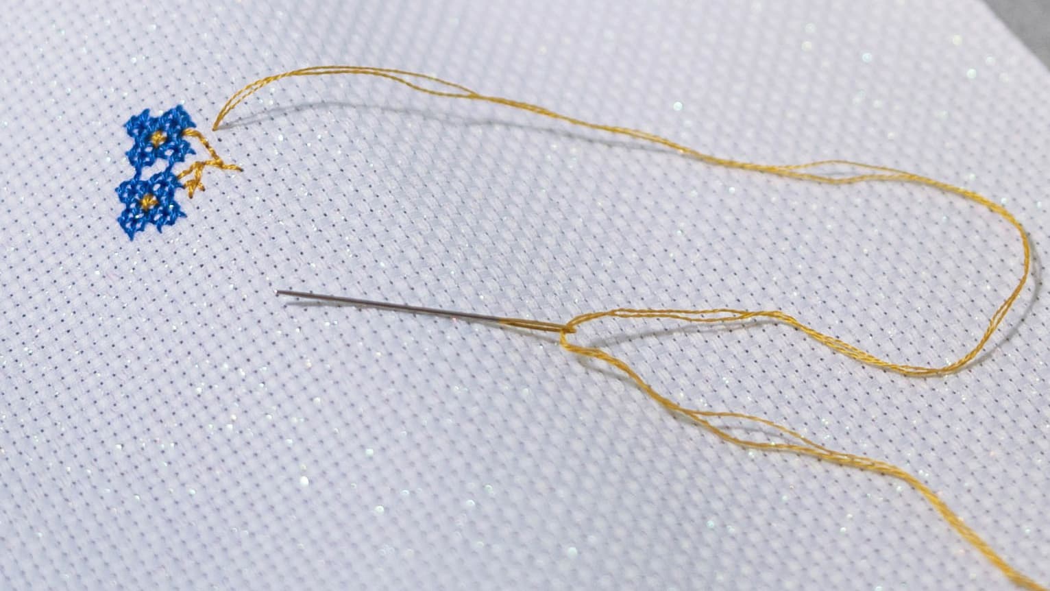 Cross-stitch needle and golden thread on white Aida cloth with small blue floral stitches