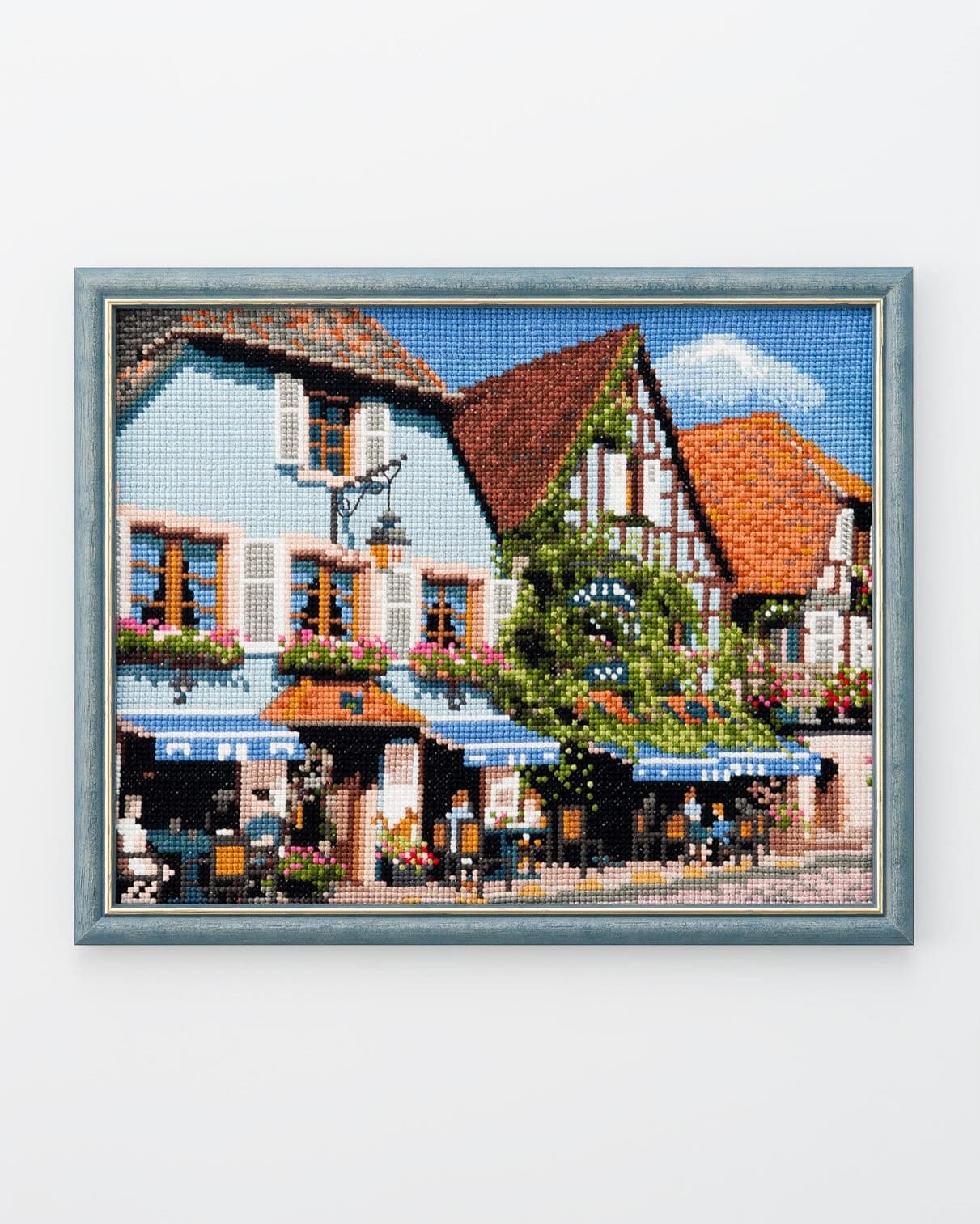 Framed pictorial cross-stitch of a European café street with blue shutters, flower boxes, and terracotta roofs