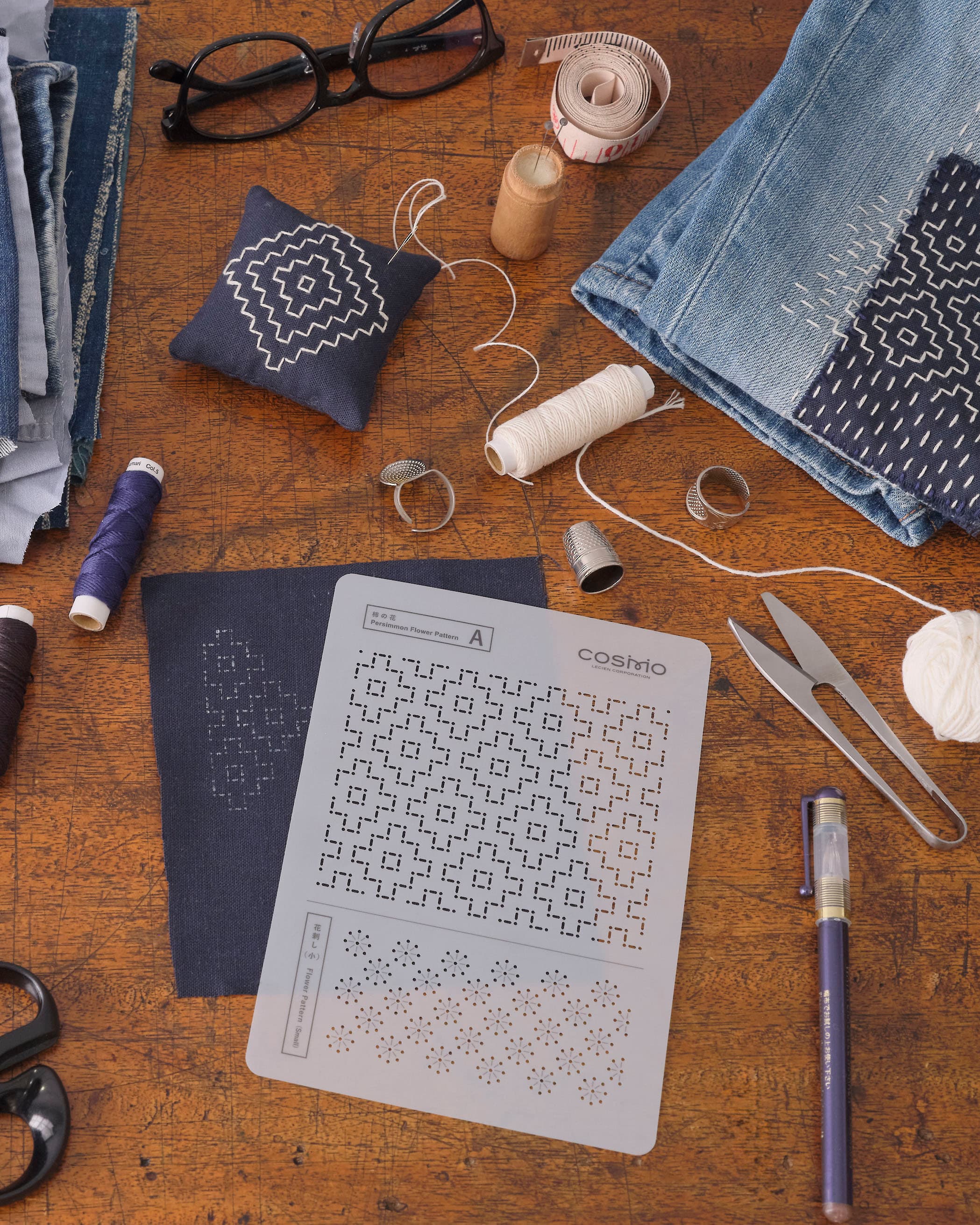 Indigo sashiko fabric with white geometric stitching, sashiko needle, and COSMO thread on a wooden work surface
