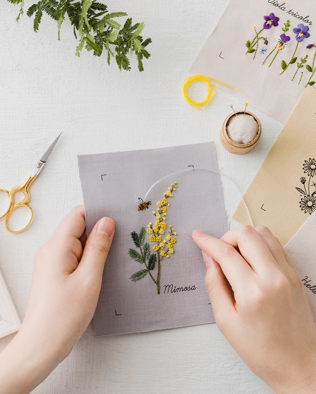 Hands stitching a mimosa embroidery with yellow French knots, scissors, and botanical pieces nearby