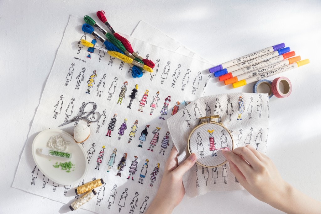 Hands stitching a 100 Ladies figure in a hoop, surrounded by COSMO embroidery floss, fabric markers, and beads