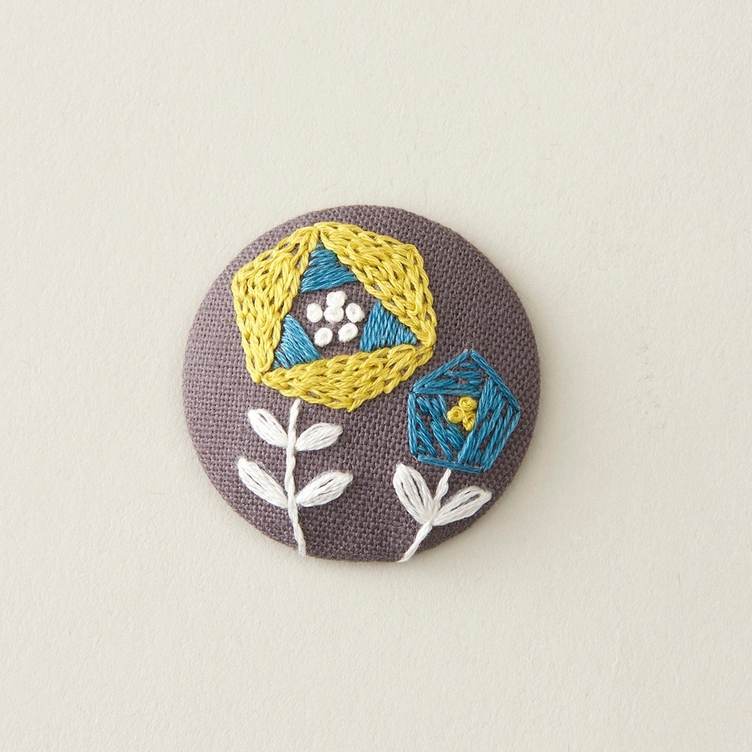 Circular brooch with yellow and teal geometric flower embroidery on dark purple cloth