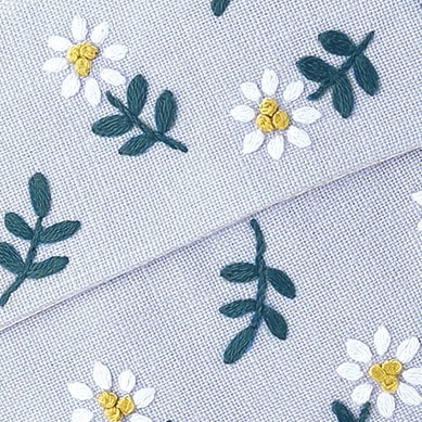 Close-up of white daisy embroidery with yellow French-knot centers and green leaves on light blue linen
