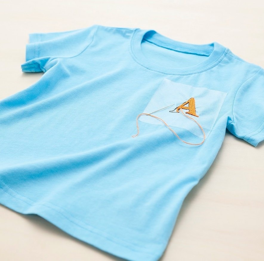 Light blue children’s tee with orange satin-stitch letter A in progress on the chest pocket