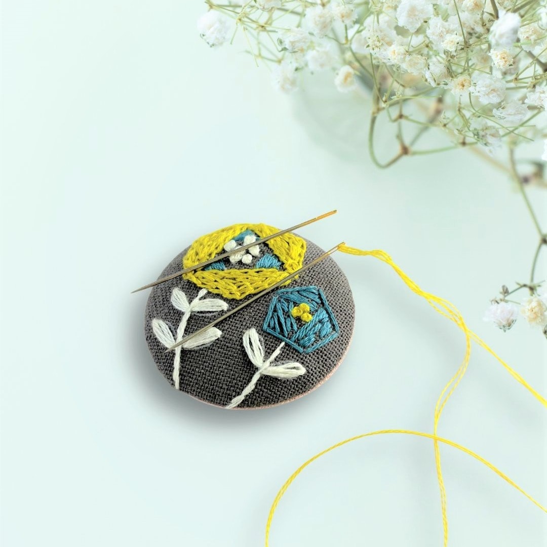 Embroidered floral brooch in progress with yellow thread and needles on grey fabric