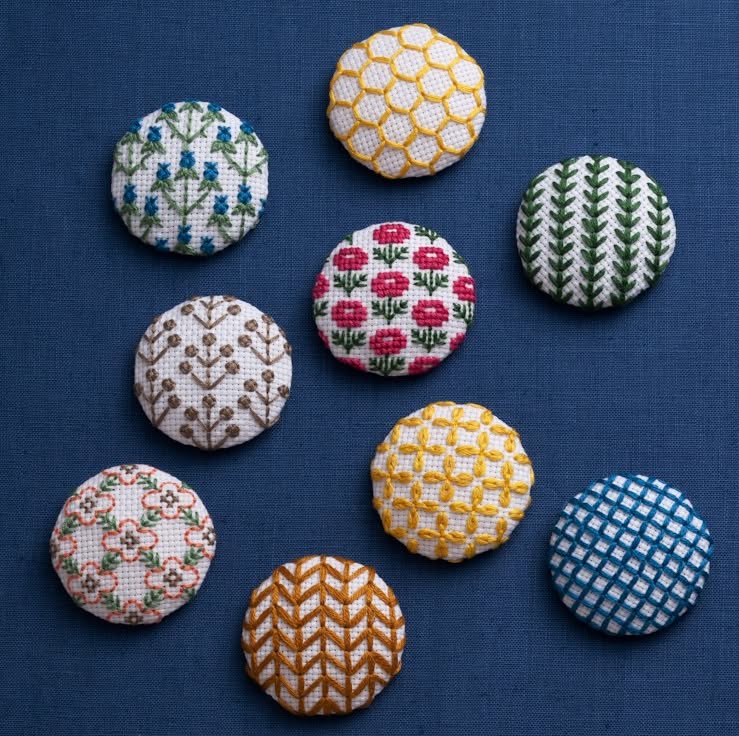 Nine embroidered covered buttons with floral and geometric motifs on blue cloth