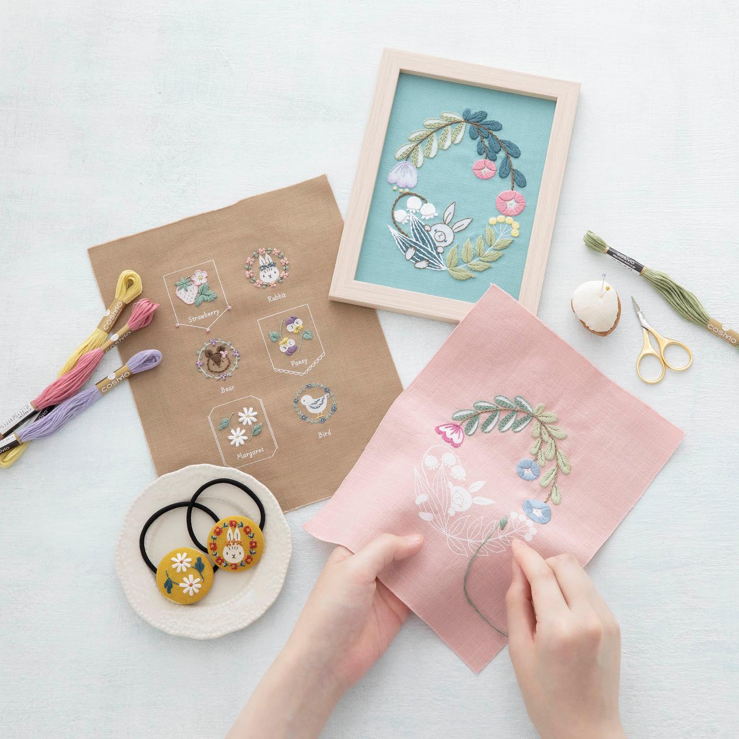 Hands stitching a floral wreath beside a finished framed bunny embroidery