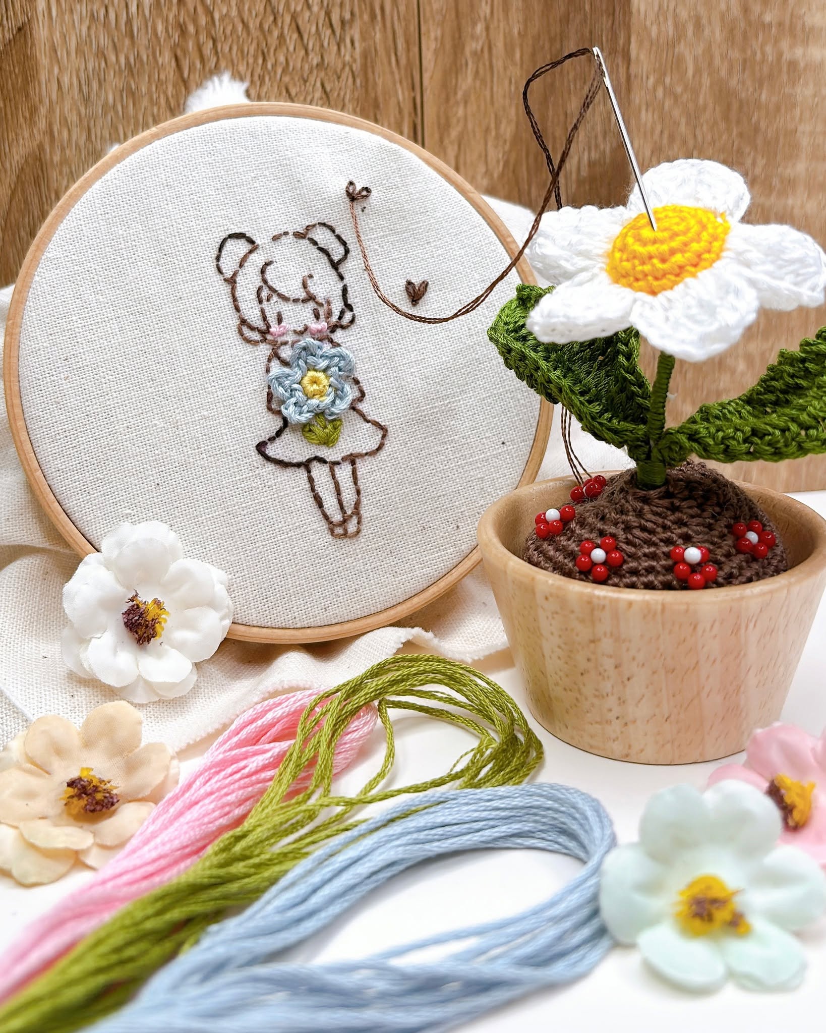 Embroidery hoop with line-art girl holding a crochet flower beside a crochet daisy in a wooden pot