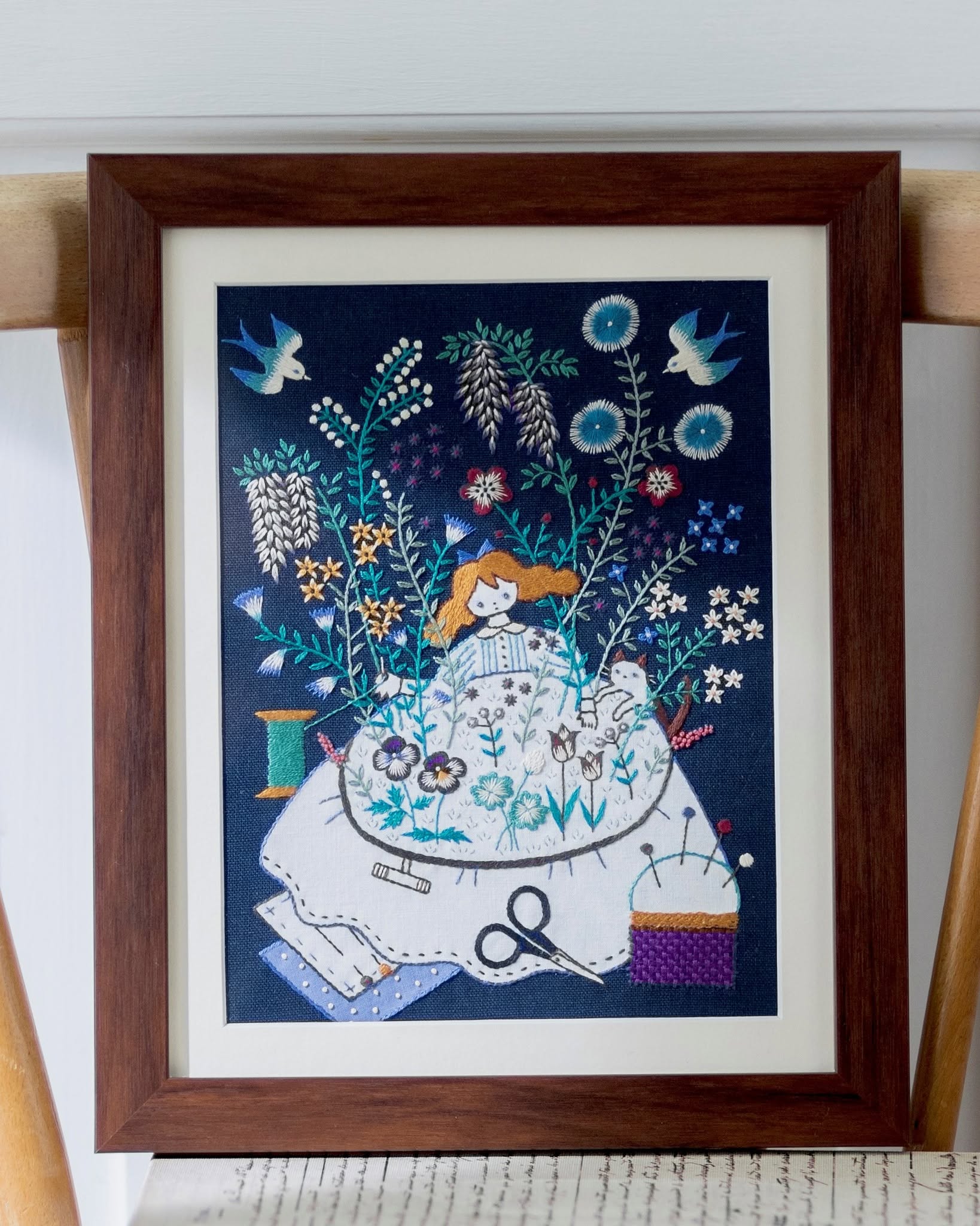 Framed embroidery of a girl stitching pansies among garden flowers and birds on navy fabric