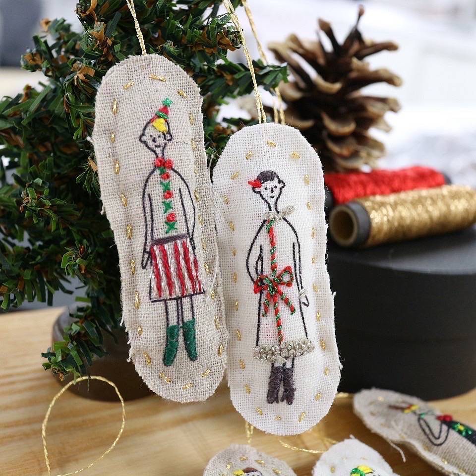 Two embroidered holiday ornaments hanging from a small evergreen beside metallic thread