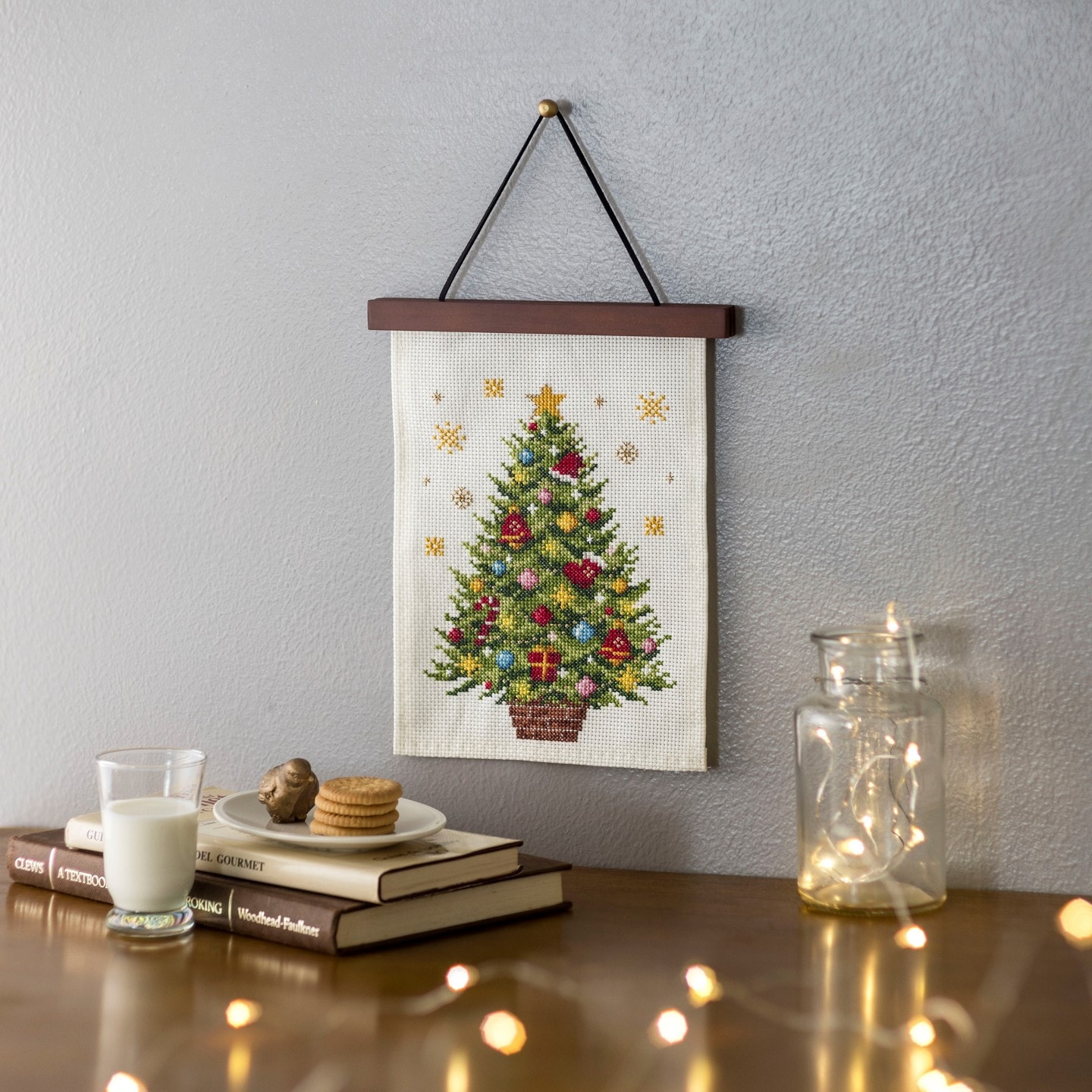 Cross-stitch Christmas tree wall hanging above books, milk, and fairy lights