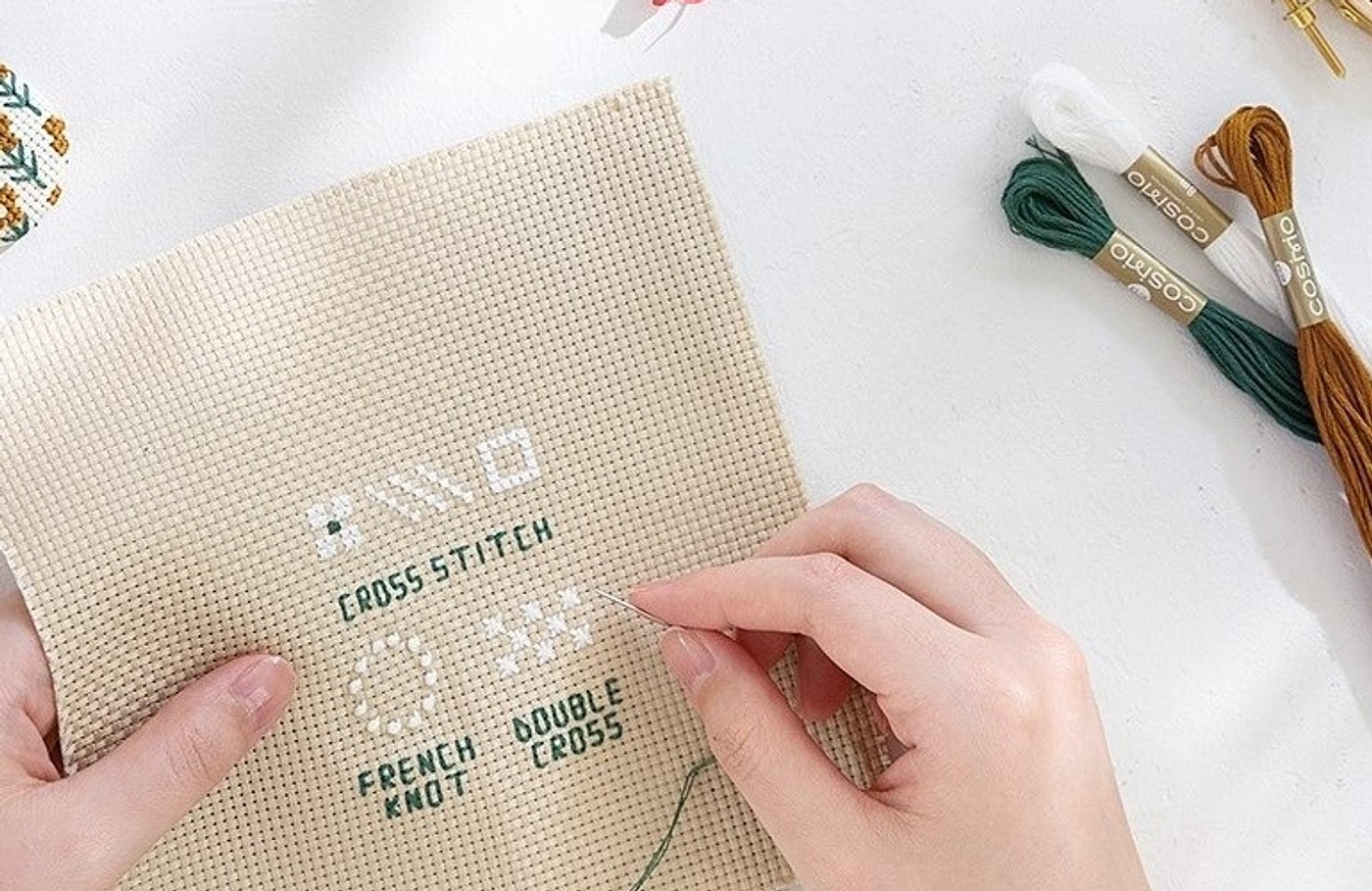 Hands cross-stitching a sampler on cream Aida cloth with COSMO floss
