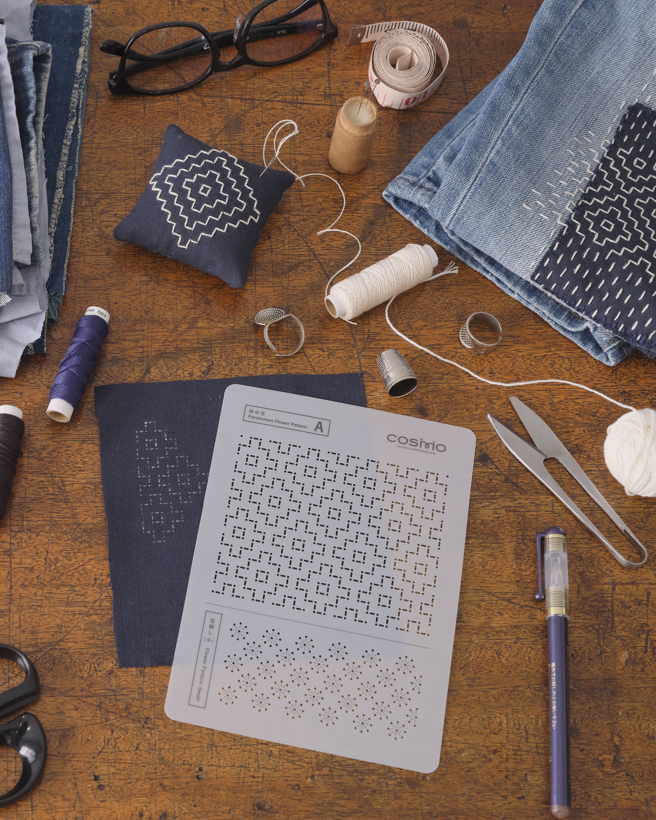 Indigo sashiko fabric with white geometric stitching, sashiko needle, and COSMO thread on a wooden work surface