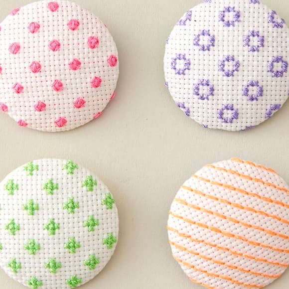 Four covered buttons with fluorescent cross-stitch patterns in pink, purple, green, and orange Neoni thread