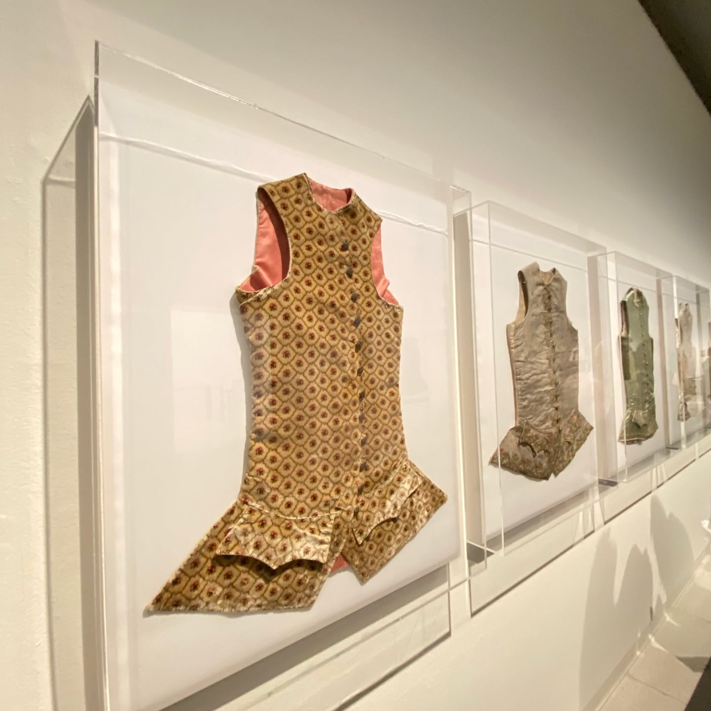 Four historical waistcoats displayed in clear wall cases, showing how embroidered and patterned panels read as complete garment faces
