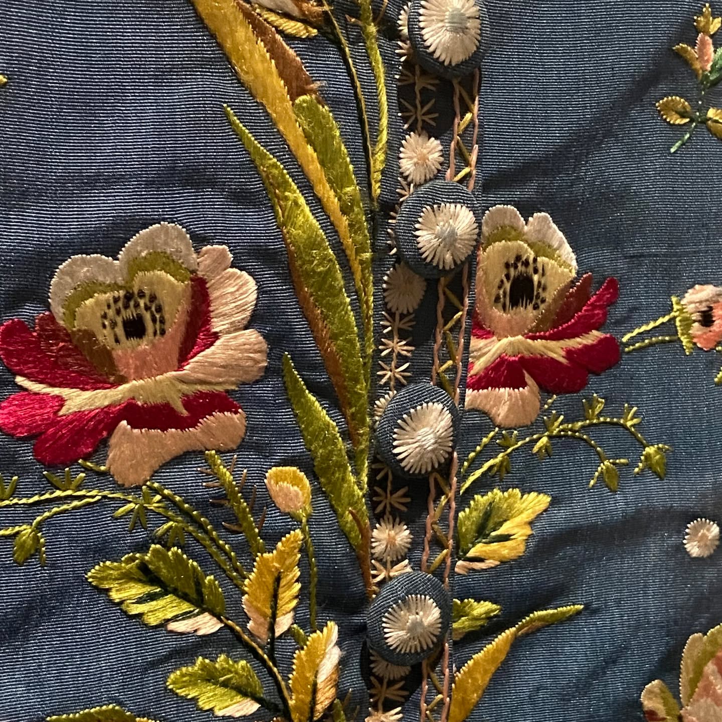 Detail of magenta-to-cream embroidered roses and yellow-green leaves on dark teal ribbed cloth beside sunburst buttons