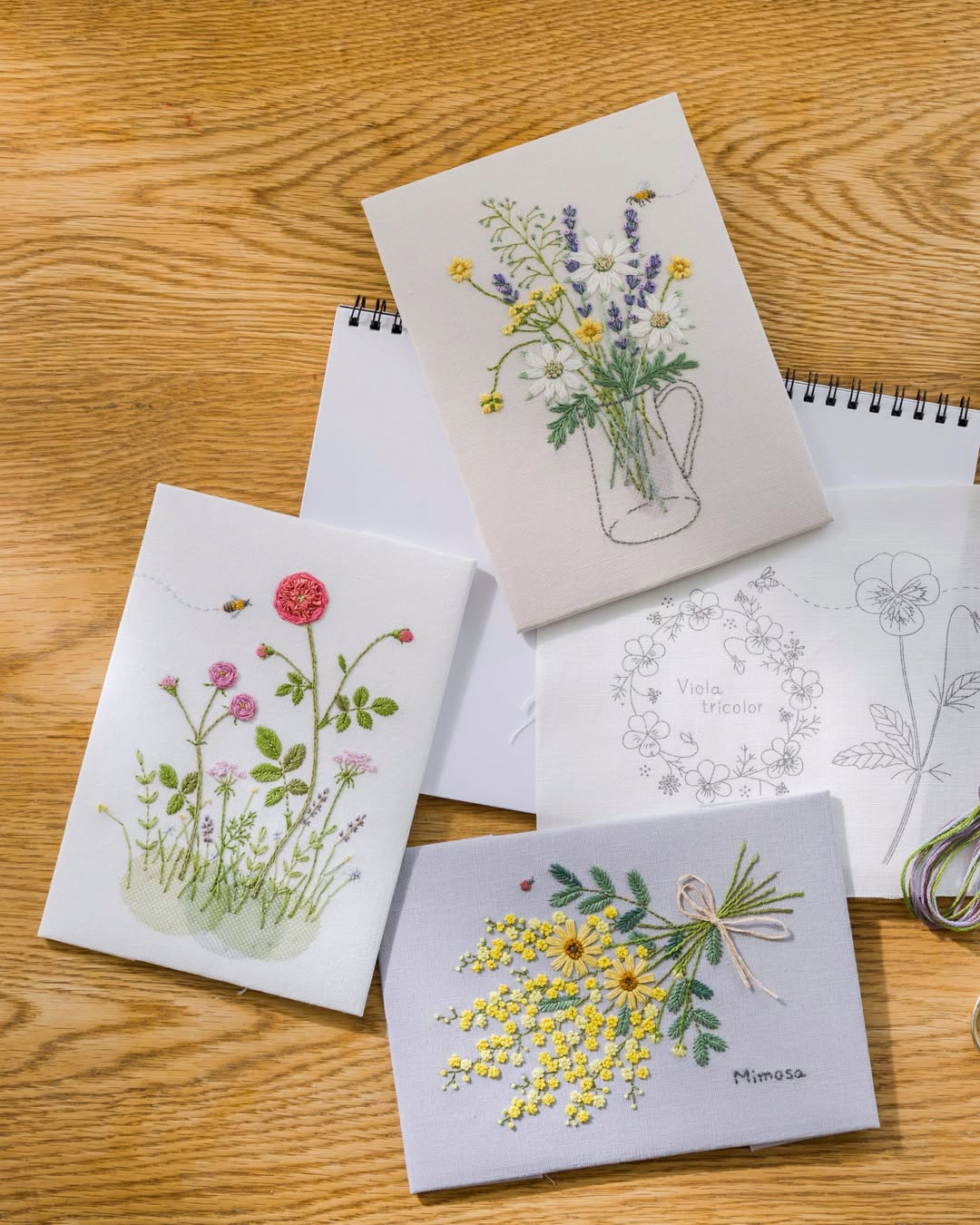 Finished floral embroidery boards and a work-in-progress with a loop of variegated thread on a wooden table
