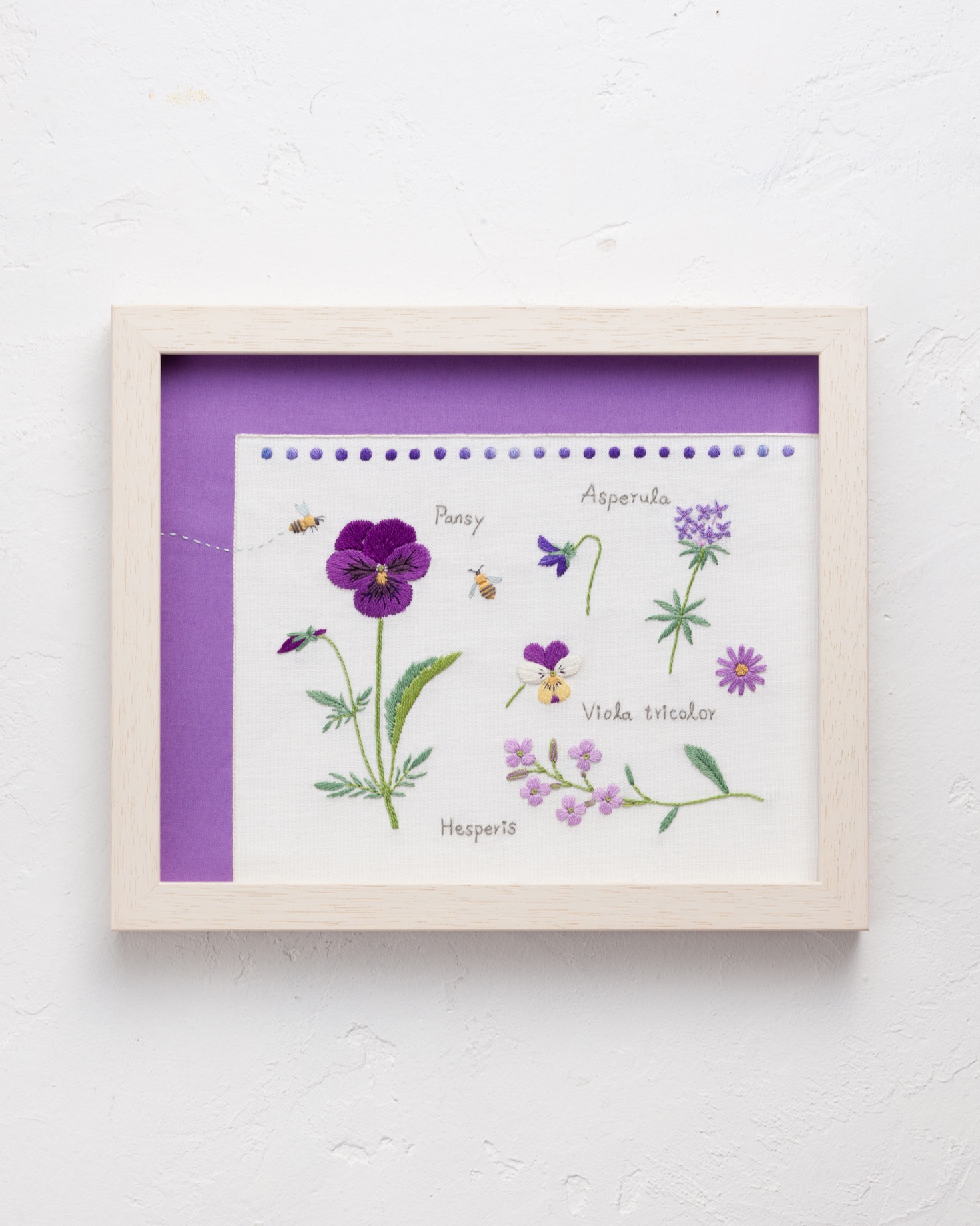 Framed botanical embroidery of pansy, viola, asperula, and hesperis with small stitched bees